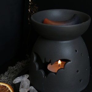 May include: Black ceramic wax warmer with a bat design and a small white candle burning inside. The warmer has a small dish on top with two wax melts, one orange and one blue.