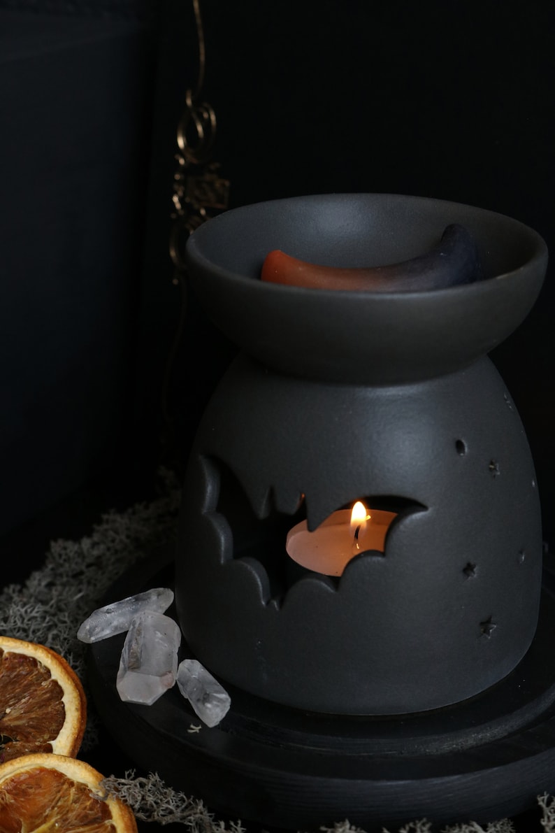 May include: Black ceramic wax warmer with a bat design and a small white candle burning inside. The warmer has a small dish on top with two wax melts, one orange and one blue.