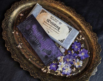 May include: Two scented wax bars from Wolf Witch Apothecary, one purple and one gray, rest on a decorative gold tray with purple flowers. The gray bar is labeled "Wildling" and the purple bar says "Wolf Witch Apothecary".