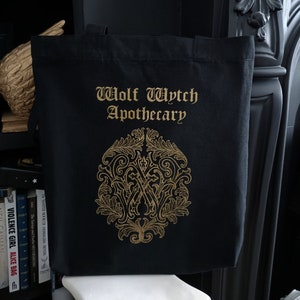 May include: Black canvas tote bag with gold lettering that reads "Wolf Wytch Apothecary". A gold ornate design with a "W" in the center is below the text.