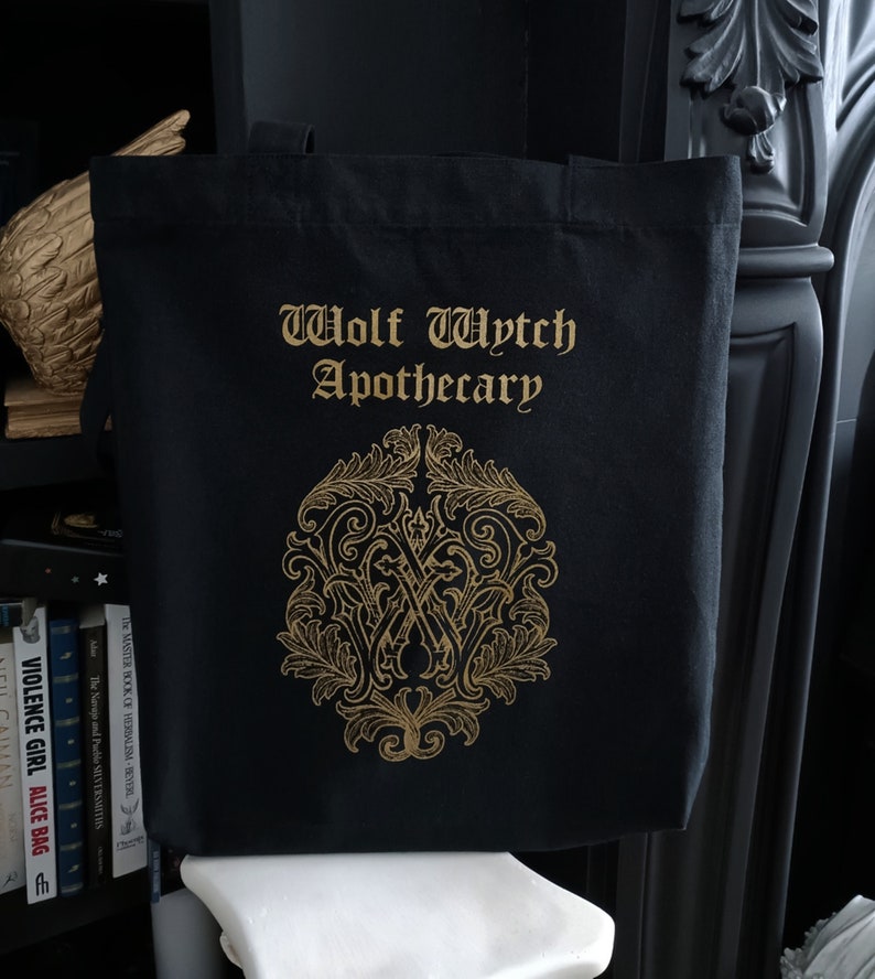 May include: Black canvas tote bag with gold lettering that reads "Wolf Wytch Apothecary". A gold ornate design with a "W" in the center is below the text.