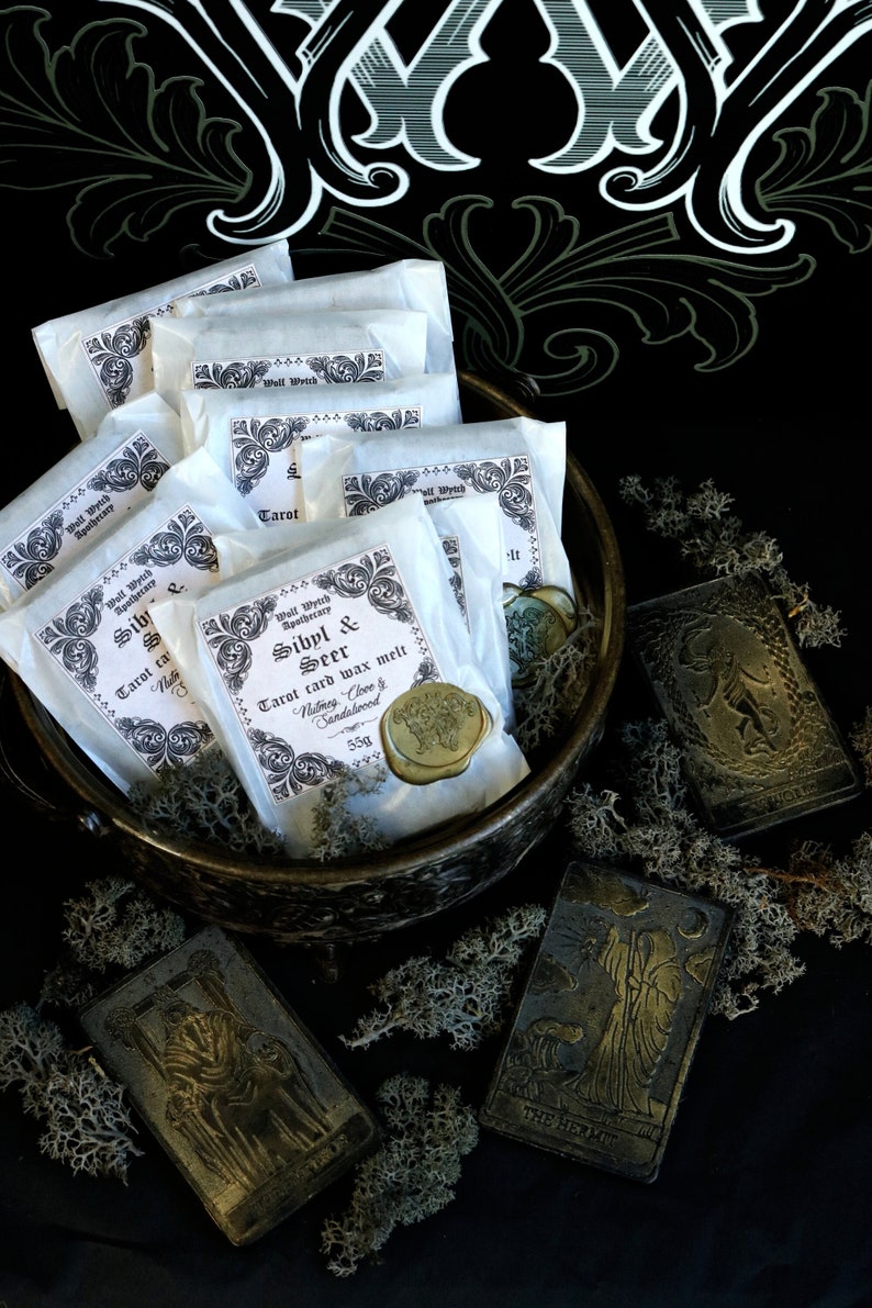 May include: A close-up of a metal bowl filled with white paper packets labeled "Sibyl & Seer Tarot card wax melt" with a gold wax seal. The bowl is surrounded by black and gold tarot cards, including "The Hermit".