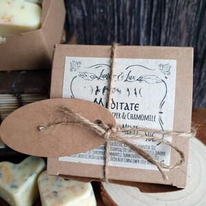 May include: A close-up of a brown box tied with twine, labeled "Meditate" with "Juniper & Chamomile" text. Heart-shaped wax melts with floral inclusions are visible. The product is on a wooden surface.