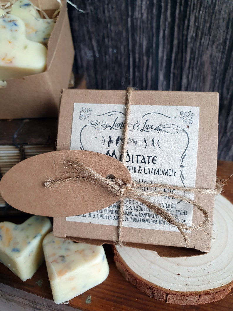May include: A close-up of a brown box tied with twine, labeled "Meditate" with "Juniper & Chamomile" text. Heart-shaped wax melts with floral inclusions are visible. The product is on a wooden surface.