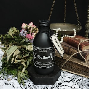 Pode incluir: Uma garrafa preta de recarga de difusor de palheta com um rótulo que diz "Hallow Tobacco & Dark Honey". A garrafa está em um suporte preto, com flores secas e livros antigos no fundo. A garrafa contém 100 ml de líquido.