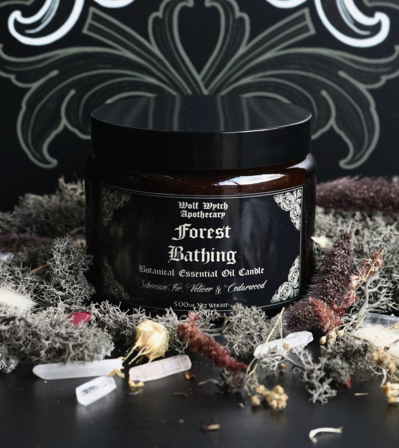 K&ouml;nnte beinhalten: Ein braunes Glasgef&auml;&szlig; mit schwarzem Deckel, beschriftet mit "Wolf Wytch Apothecary Forest Bathing Botanical Essential Oil Candle Siberian Fir Vetiver & Cedarwood 500ml NET WEIGHT". Das Gef&auml;&szlig; ist von Moos, Kristallen und getrockneten Blumen umgeben.