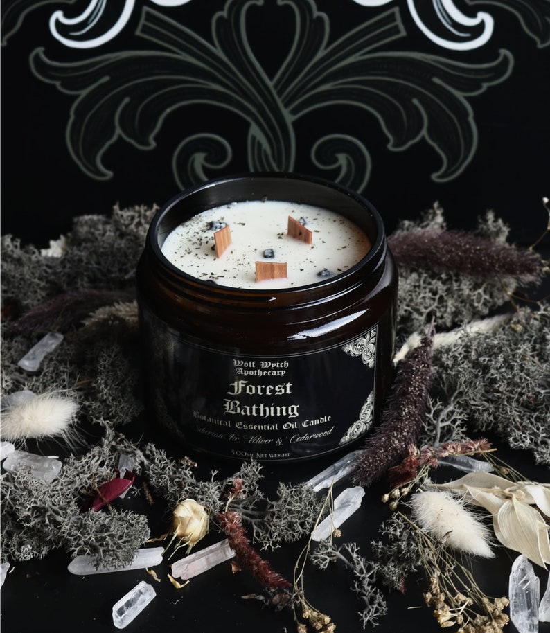 K&ouml;nnte beinhalten: Ein braunes Glasgef&auml;&szlig; mit einer Kerze, die ein schwarzes Etikett mit der Aufschrift "Wolf Witch Apothecary Forest Bathing Botanical Essential Oil Candle Scents: Fir, Vetiver & Cedarwood 5.0 oz Net Weight" tr&auml;gt. Die Kerze ist angez&uuml;ndet und hat eine wei&szlig;e Wachsoberfl&auml;che mit Holzst&auml;bchen und schwarzen Kristallen, die in das Wachs eingebettet sind.