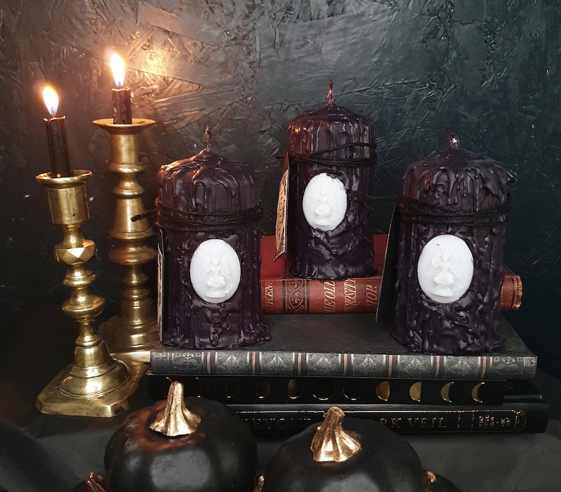 May include: Four black pillar candles with white wax medallions on a stack of books. The candles are on a dark surface with two gold candle holders.