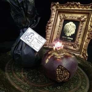 May include: A dark purple pomegranate-shaped candle with gold accents sits on a decorative tray. The candle is lit and has a small flame. The candle is labelled "Persephone's Fruit" and "Black Witch Botanicals".