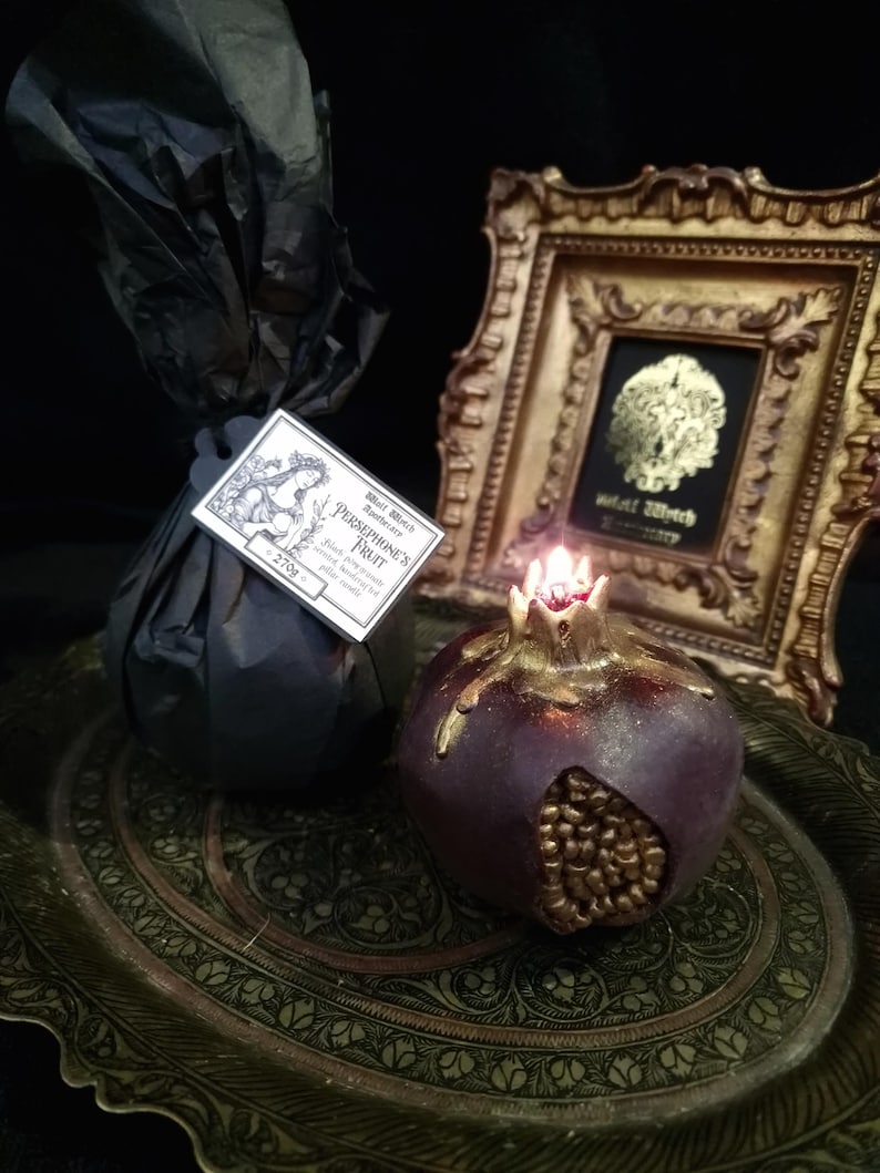 May include: A dark purple pomegranate-shaped candle with gold accents sits on a decorative tray. The candle is lit and has a small flame. The candle is labelled "Persephone's Fruit" and "Black Witch Botanicals".
