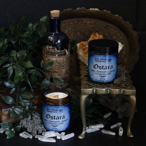 May include: Two amber glass candle jars with blue labels that read "Wolf Witch Apothecary Ostara Spruce, Peppermint & Jasmine Coconut wax candle 180h Net Weight 11oz" on a dark surface with a gold metal stand, green foliage, and clear quartz crystals.