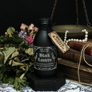 Puede incluir: Botella de vidrio negro con una etiqueta negra que dice "Wolf Witch Apothecary Black Lantern Incense, Leather & Sandalwood Luxury Reed Diffuser Refill 100ML". La botella está sobre una base hexagonal negra.