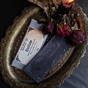 May include: A still-life arrangement featuring two bars of soap from Wolf Wytch Apothecary, one labeled "Hallow" and the other with the brand name. The soaps rest on an ornate gold-colored tray, accompanied by dried roses and a lit black candle.