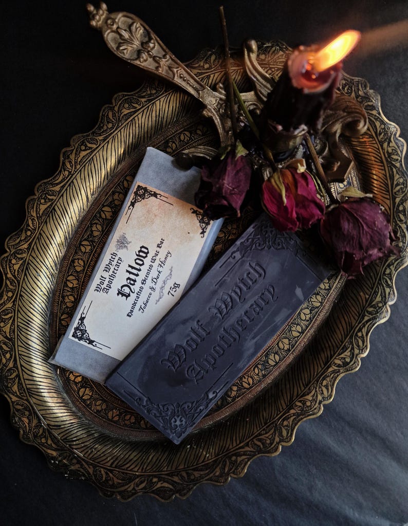 May include: A still-life arrangement featuring two bars of soap from Wolf Wytch Apothecary, one labeled "Hallow" and the other with the brand name. The soaps rest on an ornate gold-colored tray, accompanied by dried roses and a lit black candle.
