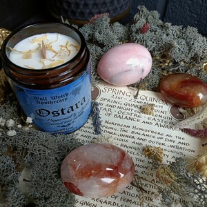 May include: A candle in a glass jar with a blue label that reads "Wolf Wytch Apothecary Ostara Peppermint & Coconut  Incense". The candle is surrounded by pink, red, and orange stones and a piece of paper with text about the Spring Equinox.