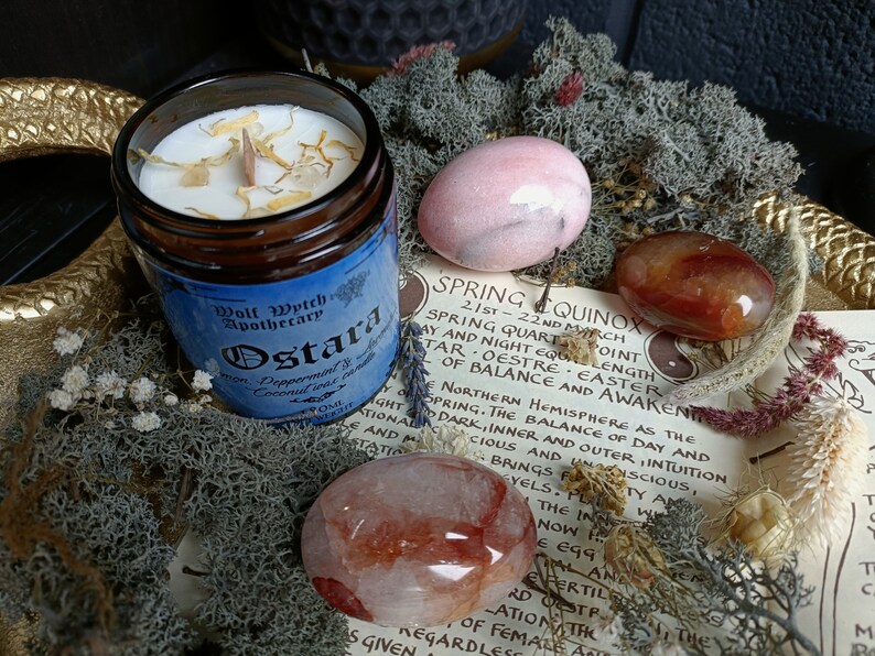 May include: A candle in a glass jar with a blue label that reads "Wolf Wytch Apothecary Ostara Peppermint & Coconut  Incense". The candle is surrounded by pink, red, and orange stones and a piece of paper with text about the Spring Equinox.