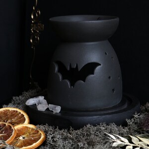 May include: Black ceramic wax warmer with a bat cutout design. The warmer is on a wooden base and surrounded by dried orange slices, crystals, and moss.