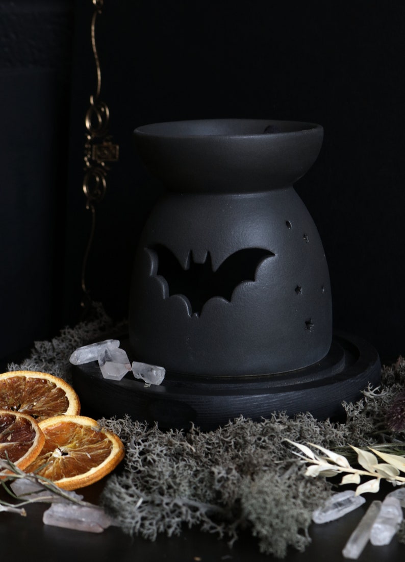 May include: Black ceramic wax warmer with a bat cutout design. The warmer is on a wooden base and surrounded by dried orange slices, crystals, and moss.