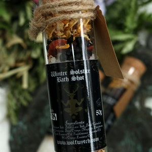 May include: A glass tube filled with bath salts, dried orange peel, and other herbs. The tube has a black label that reads "Winter Solstice Bath Shot" and includes a list of ingredients. The label also includes the website address "www.wolfwytch.com".