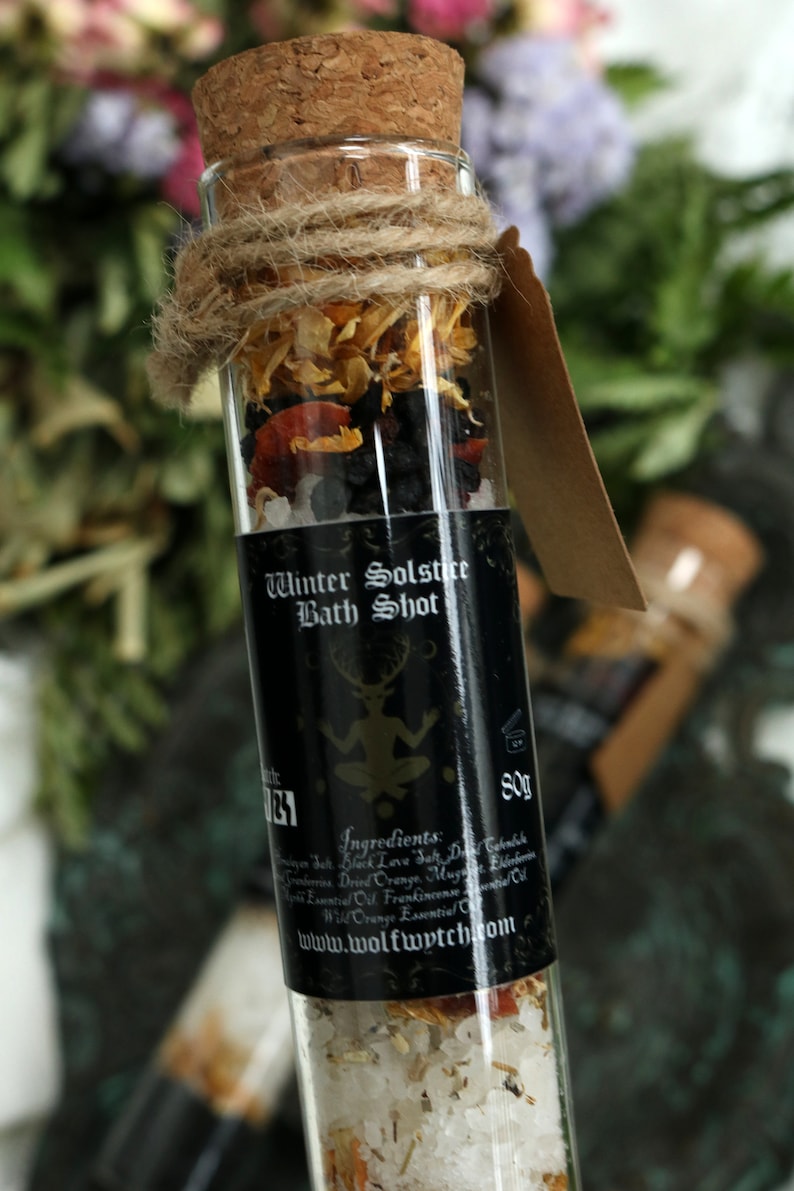 May include: A glass tube filled with bath salts, dried orange peel, and other herbs. The tube has a black label that reads "Winter Solstice Bath Shot" and includes a list of ingredients. The label also includes the website address "www.wolfwytch.com".