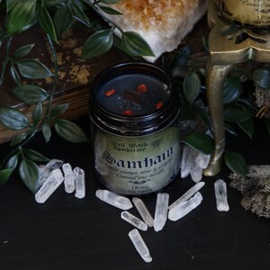 May include: A black candle jar with a green label that reads "Wolf Watch Apothecary Samhain Wild orange clove & ginger Coconut wax candle 180ML NET WEIGHT". The candle jar is sitting on a black surface with clear quartz crystals scattered around it.