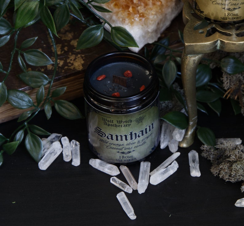 May include: A black candle jar with a green label that reads "Wolf Watch Apothecary Samhain Wild orange clove & ginger Coconut wax candle 180ML NET WEIGHT". The candle jar is sitting on a black surface with clear quartz crystals scattered around it.