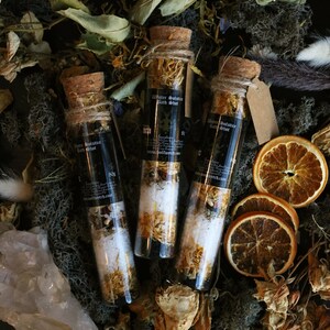 May include: Three small glass bottles filled with bath salts and dried flowers. The bottles have cork stoppers and are labeled "Winter Solstice Bath Shot".