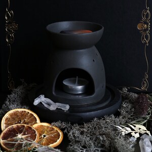 May include: Black ceramic oil diffuser with a round opening for a tea light candle. The diffuser is sitting on a black wooden base with a white crystal on top. The diffuser is surrounded by dried orange slices, moss, and other natural elements.