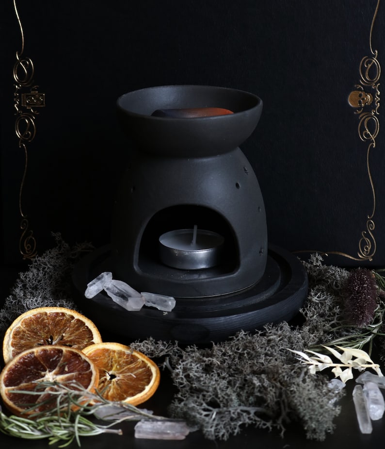 May include: Black ceramic oil diffuser with a round opening for a tea light candle. The diffuser is sitting on a black wooden base with a white crystal on top. The diffuser is surrounded by dried orange slices, moss, and other natural elements.