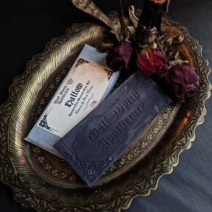 May include: A dark, moody still life featuring two bars of soap from Wolf Witch Apothecary on an ornate, gold-toned tray. One bar is wrapped in paper, the other is dark gray with embossed text. A lit black candle and dried roses add to the gothic aesthetic.