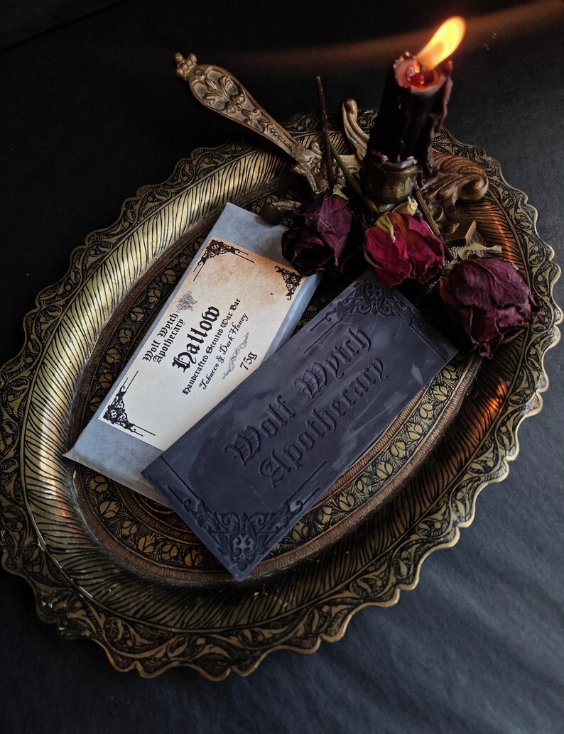 May include: A dark, moody still life featuring two bars of soap from Wolf Witch Apothecary on an ornate, gold-toned tray. One bar is wrapped in paper, the other is dark gray with embossed text. A lit black candle and dried roses add to the gothic aesthetic.