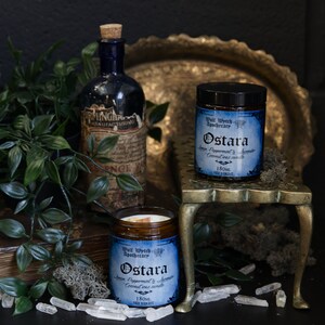 May include: Two glass jars with black lids and labels that read "Wall Watch Apothecary Ostara Ginger Peppermint & Lavender Coconut Wax Candle 180ml Net Weight". The jars are on a gold metal stand with a round top and three legs. The stand is on a dark surface with a green plant, a book, and white crystals.