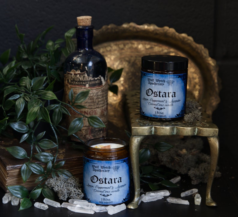 May include: Two glass jars with black lids and labels that read "Wall Watch Apothecary Ostara Ginger Peppermint & Lavender Coconut Wax Candle 180ml Net Weight". The jars are on a gold metal stand with a round top and three legs. The stand is on a dark surface with a green plant, a book, and white crystals.