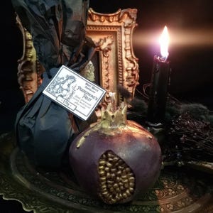May include: A black and gold pomegranate-shaped candle with the text "Wild Witch Apothecary Persephone's Fruit Black pomegranate scent, hand-poured, palm wax candle" on a label. The candle is sitting on a gold tray with a black candle in the background.