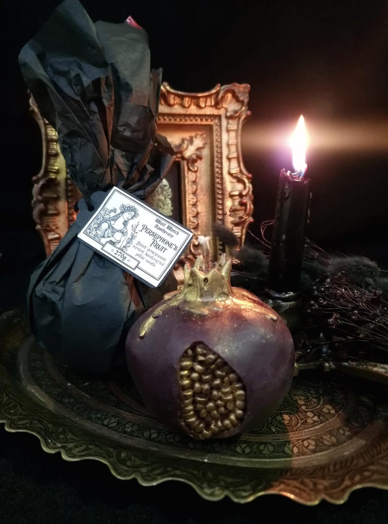 May include: A black and gold pomegranate-shaped candle with the text "Wild Witch Apothecary Persephone's Fruit Black pomegranate scent, hand-poured, palm wax candle" on a label. The candle is sitting on a gold tray with a black candle in the background.