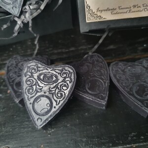May include: Four black heart-shaped wax melts with a silver design of an eye and a crescent moon. The wax melts are on a dark wooden surface.
