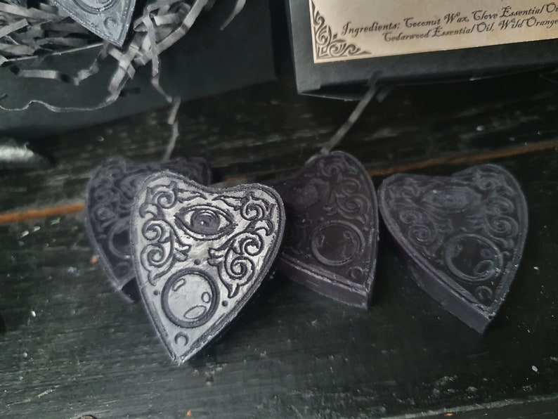 May include: Four black heart-shaped wax melts with a silver design of an eye and a crescent moon. The wax melts are on a dark wooden surface.
