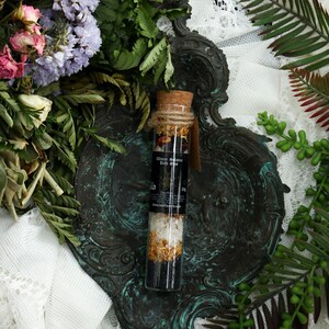 May include: A glass bottle filled with bath salts and dried flowers, with a cork stopper and a black label that reads "Water Wanderer Bath Soak". The bottle is sitting on a decorative, ornate, dark green metal tray.