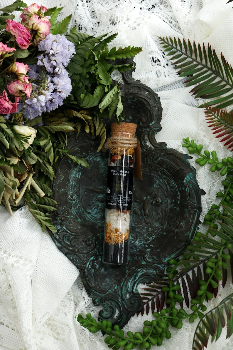 May include: A glass bottle filled with bath salts and dried flowers, with a cork stopper and a black label that reads "Water Wanderer Bath Soak". The bottle is sitting on a decorative, ornate, dark green metal tray.
