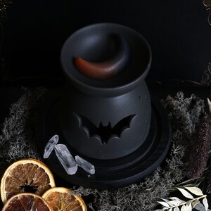 May include: Black ceramic oil burner with a crescent moon design and a bat cutout. The burner is on a black wooden base and surrounded by crystals, dried orange slices, and dried foliage.