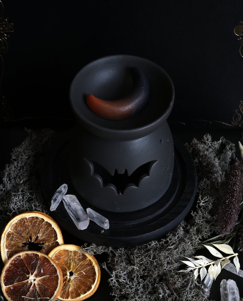 May include: Black ceramic oil burner with a crescent moon design and a bat cutout. The burner is on a black wooden base and surrounded by crystals, dried orange slices, and dried foliage.