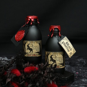 May include: Two black glass bottles with red wax seals and labels. The labels feature a black goat illustration and text that reads "Wall Witch Apothecary", "Delicately Dark Oud & Spicy Cherry", "Luxury Reed Diffuser", "200ml", "Black Phillip" and "Black Phillip II".