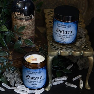 May include: Two amber glass jars with blue labels and black lids. The labels read "Wolf Wytch Apothecary" and "Ostara Lemon, Peppermint & Lavender Coconut Wax Candle 180ml Net Weight". The jars are on a gold metal stand with a wooden base. The stand is surrounded by clear quartz crystals.