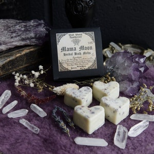 Op de afbeelding: Vier witte hartvormige badmelts met lavendel en gedroogde bloemen, verpakt in een zwarte doos met de tekst "Mama Moon Herbal Bath Melts" en "Wolf Witch Apothecary". De badmelts worden gepresenteerd op een paarse fluwelen ondergrond met heldere kwartskristallen en amethist.