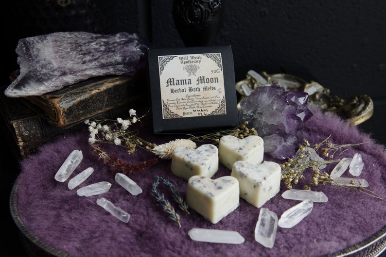 Op de afbeelding: Vier witte hartvormige badmelts met lavendel en gedroogde bloemen, verpakt in een zwarte doos met de tekst "Mama Moon Herbal Bath Melts" en "Wolf Witch Apothecary". De badmelts worden gepresenteerd op een paarse fluwelen ondergrond met heldere kwartskristallen en amethist.