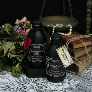 Könnte beinhalten: Zwei schwarze Apothekerflaschen mit Etiketten "Persephone's Fruit" und "Black Pomegranate", je 100 ml. Eine Flasche hat ein rotes Wachssiegel. Der Hintergrund umfasst getrocknete Blumen, alte Bücher und eine Waage.