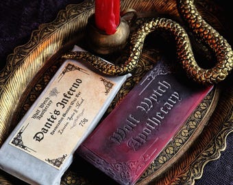 May include: A still-life arrangement featuring two scented wax bars from Wolf Wytch Apothecary, one wrapped in paper and the other in a deep red hue. A lit red candle sits in a brass holder, and a golden snake winds around the bars on a decorative tray.