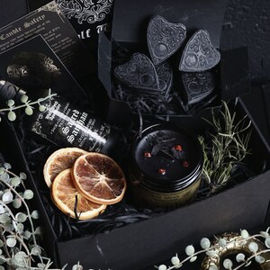 May include: A black gift box filled with various items, including a black candle, dried orange slices, a bottle of sage and clove oil, and black heart-shaped charms. The box is surrounded by green leafy sprigs.
