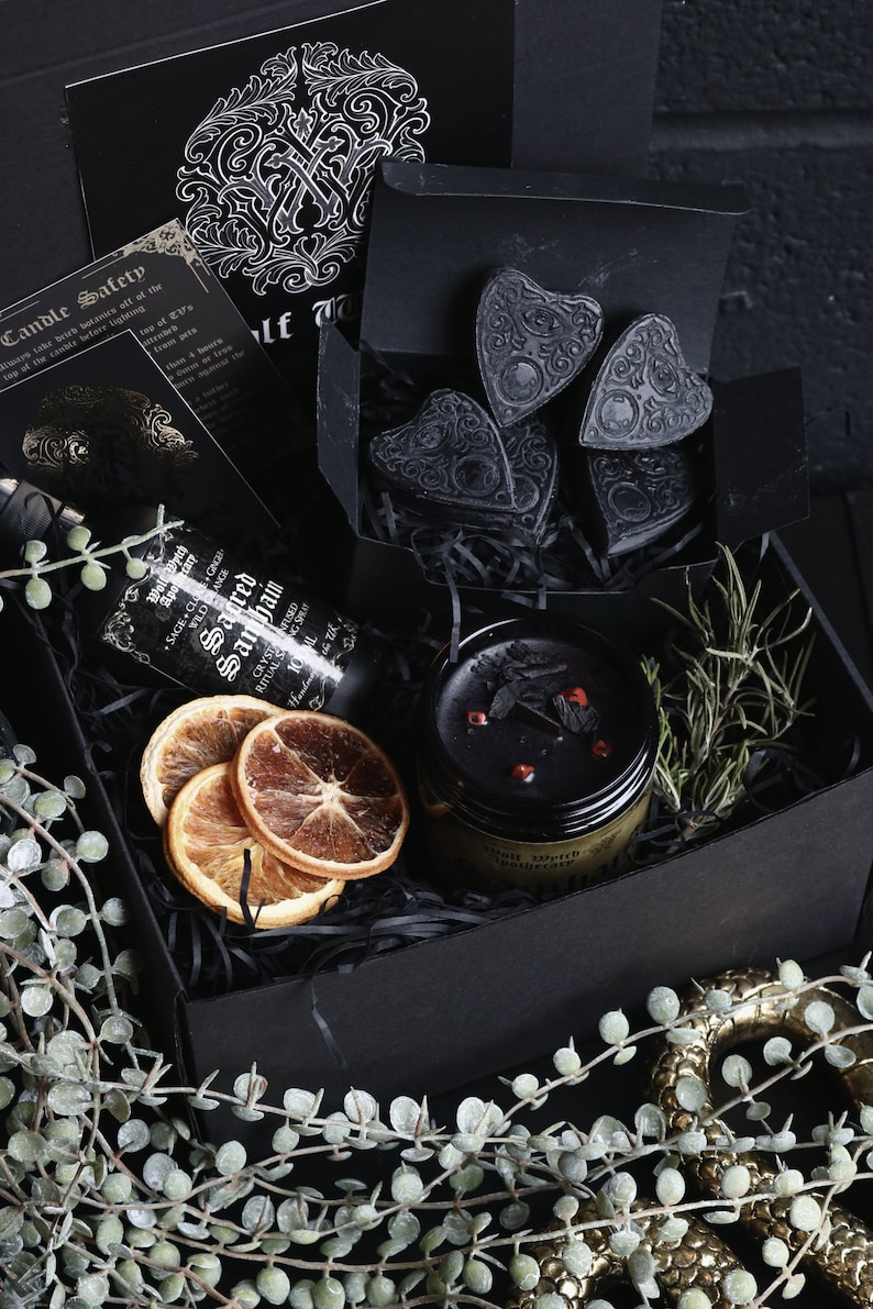 May include: A black gift box filled with various items, including a black candle, dried orange slices, a bottle of sage and clove oil, and black heart-shaped charms. The box is surrounded by green leafy sprigs.