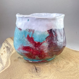 Könnte beinhalten: Eine handgefertigte Keramikschale mit einer einzigartigen, strukturierten Glasur. Die Schale hat einen weißen Rand, der in eine türkisfarbene und rote Mitte übergeht, und einen dunkelbraunen Boden. Die Schale steht auf einer Holzoberfläche.